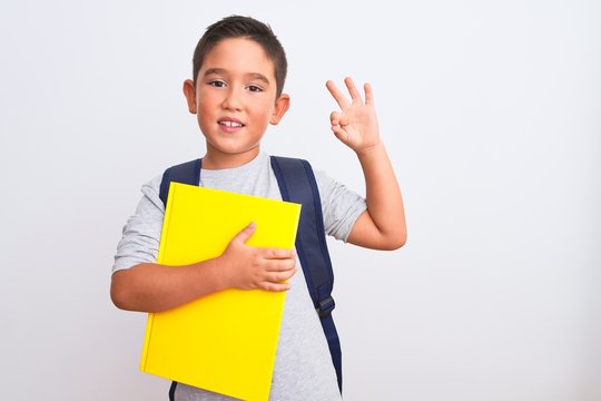Beautiful student kid boy wearing backpack holding book over isolated white background doing ok sign with fingers, excellent symbol