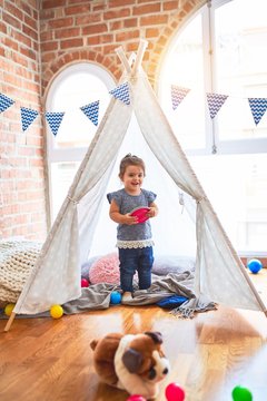 Beautiful toddler standing inside tipi holding plastic dishes smiling at kindergarten
