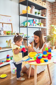 Young beautiful teacher and toddler playing with dishes, cutlery and cups toy on the table at kindergarten