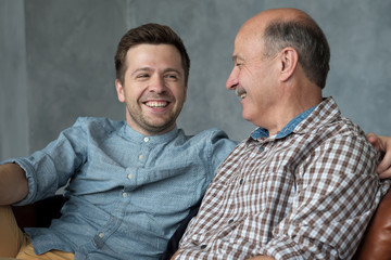 Smiling senior father sit on couch talk chat with millennial son spend time at home together