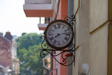 Fototapeta na wymiar Old clock on the wall. Street clock in ornate forging. Old-fashioned clock on the wall of building, close up