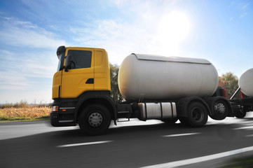 Big metal fuel tanker truck shipping fuel on the countryside road in motion against sky with bright sun