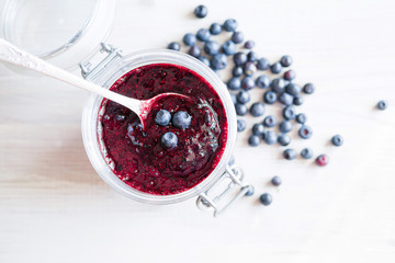 Fresh forest blueberries and blueberries jam in a glass jar on a wooden white surface, top view