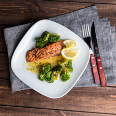 Grilled trout steak on a white plate with broccoli and lime. Cutlery on the wooden background