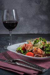 Salad with prosciutto and parmesan. Cutlery, knives, forks and pink napkin on the grey background. Glass of red wine