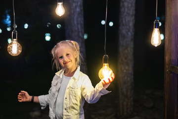 Cute adorable caucasian blond girl portrait smiling and holding in hand one of hanged edison light...