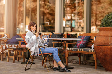 Beautiful girl resting and drinking coffee sitting  in cafe on the terrace, wrapped in a woolen plaid blanket. Autumn morning.