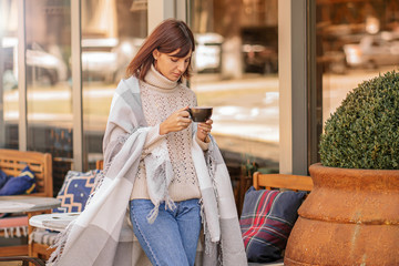 Beautiful girl resting and drinking coffee sitting  in cafe on the terrace, wrapped in a woolen plaid blanket. Autumn morning.