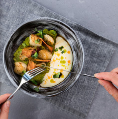 Women's hands holding spoon and fork. Scallops with mushy peas and Beurre Blanc Sauce in the Gray clay plate on a gray tablecloth