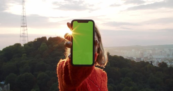Girl showing hand with mock-up smartphone chromekey greenscreen. Young woman standing at sunset in beautiful nature park with cityscape in background. Mobile gadget.