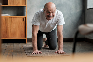 Fototapeta premium Senior hispanic man practices yoga asana marjariasana or cat-cow pose at home. Doing sport exercise for spine health.