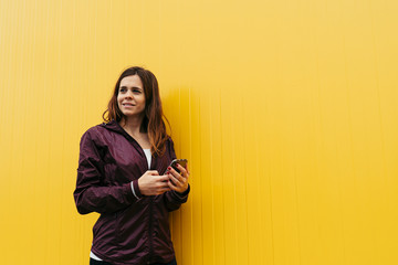 young woman with sport wear using the smartphone while is looking at the side with a yellow wall in the background © Mikel Taboada