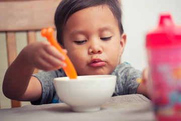 A young latino boy learning to eat independently with a spoon in hand and chewing his meal.