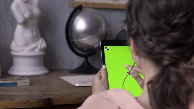 Woman hands holding tablet pc with green screen in the art studio with concrete wall and silver globe, plaster woman sculpture on the wooden table. Stock footage. Girl tapping on chromakey tablet.