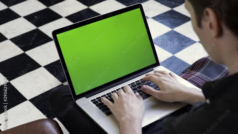 Poster over the shoulder view of a man using his computer with green screen and sitting on comfortable couc