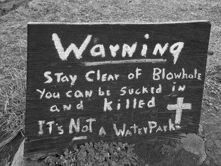Wooden sign with hand written message warning stay clear of blowhole you can be sucked in and killed it's not a waterpark in black and white