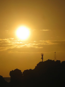 Sun setting through clouds behind silhouette of cliff diver performing daily tiki torch lighting ceremony at black rock in Maui Hawaii 