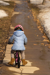 Little girl riding a balance bike on the sidewalk up a hill on a warm spring day with melting snow