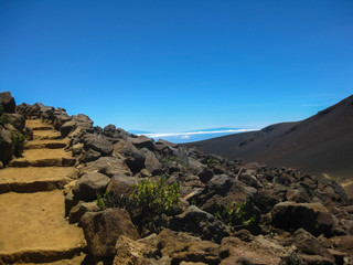 Hiking trail with stone steps along slope of dormant volcano with blue sky horizon above the clouds