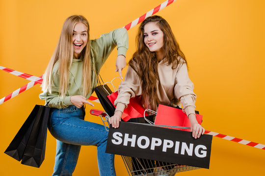 two happy smiling beautiful women have cart and shopping sign with colorful shopping bags and signal tape isolated over yellow