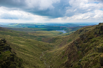 Naklejka premium Green hills that flow into a valley in the Peak District overlooking a water reservoir on a cloudy day in England, UK.