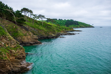 Rocky and green coastline around St Anthony Head with tropical-like waters and a lighthouse in the distance on this cloudy day in England, UK.