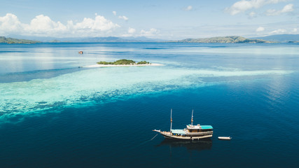 Fototapeta premium Luxury cruise boat sailing near coral reef atoll island with amazing white tropical beach and mountains on horizon. Aerial view. Luxury marine travel and vacations concept.