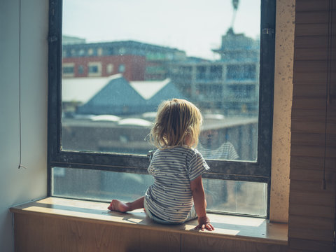 Little toddler sitting by the window in city aparetment