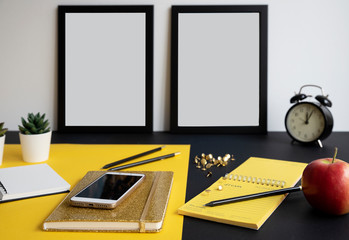 Modern office desk with blank frames with mock up. Office supplies with smartphone and gold notebook on yellow and black background. 