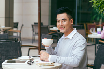 Enjoying his morning. Young Asian man holding coffee cup and looking at camera with smile while sitting in cafe - Image