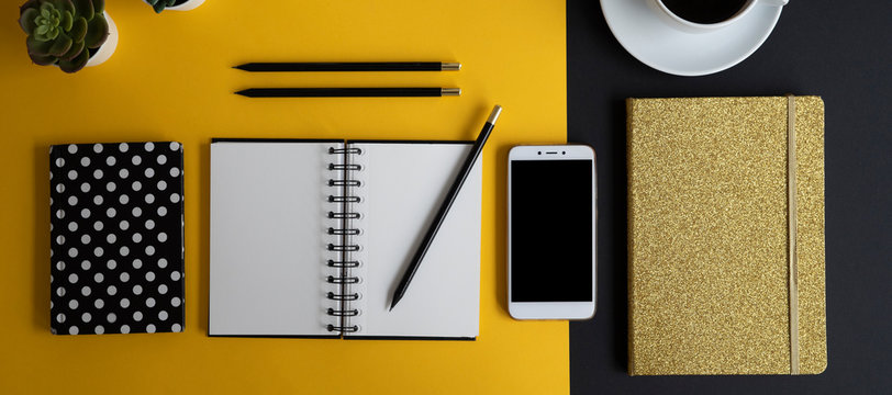 Blank notebook, smartphone, coffee and pencil on the office desk in modern design. Yellow and black create a background of desktop. Panoramic banner.