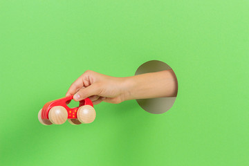 Kid holding red wooden toy car in hand through hole on light green background