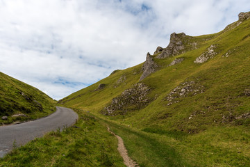 Road curves inside rocky hills covered in a green moss and grass with a small footpath adjacent to the road, on cloudy day in England, UK.