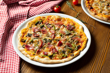Fresh homemade pizza with prosciutto, capers, olives on a wooden background in composition with a red cloth and olive oil. Italian Cuisine. Selective focus on center of pizza