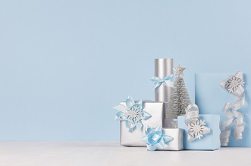 Winter festive background for advertising and design - silver glittering fir tree and gift boxes in blue, metallic color on white wood table and blue wall.