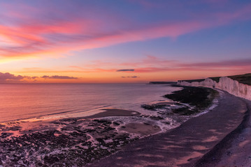 Obraz premium A sun sets over the clouds and paints golden and pink colours across the skies as the rocky shore and the white chalk cliffs of the Seven Sisters meets the sea in Eastbourne, UK.