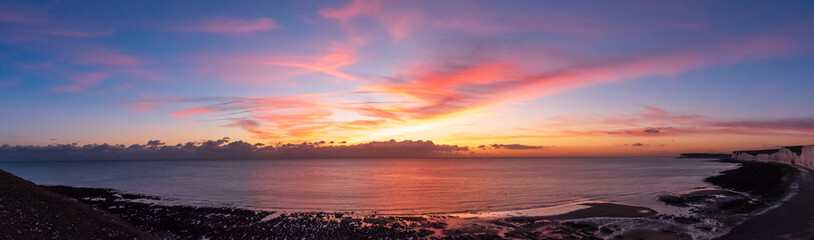 A panoramic of sun setting over the clouds and painting golden and pink colours across the skies. As the rocky shore and the white chalk cliffs of the Seven Sisters meets the sea in Eastbourne, UK.