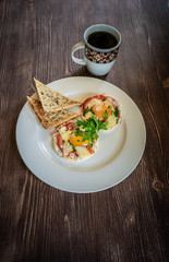 Hot breakfast on old wooden table. A pair of fried eggs with meat, vegetables and cheese on a white plate with a cup of hot coffee.