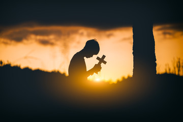 Young man sitting and kneel down for pray and worshipping God with cross under the tree at sunset background. christian silhouette concept.