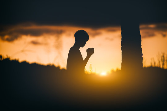 Young christian sitting and kneel down for pray under the tree at sunset background. christian silhouette concept.