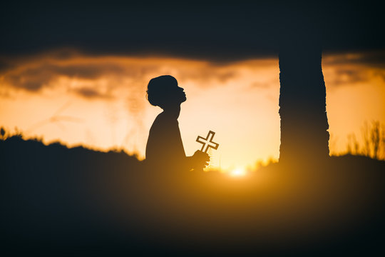 Young christian kneel down for pray and worshipping God with cross under the tree at sunset background. christian silhouette concept.