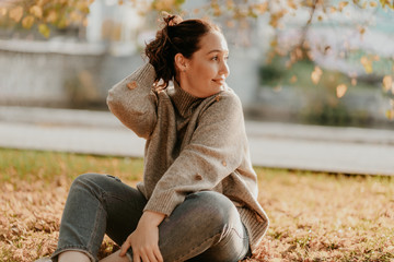 Friendly charming brunette woman with long curly hair in cozy wool sweater sitting on ground with...