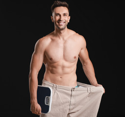Fototapeta na wymiar Handsome muscular man in loose pants and with scales on dark background. Weight loss concept