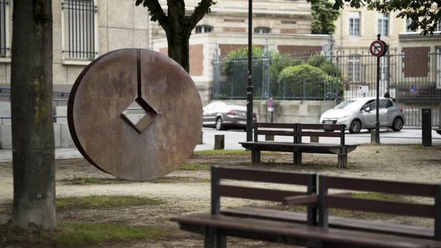 Modern city park with benches and metal round statue with a diamond-shaped hole in the middle of it. Stock footage. A public place with trees near the road.