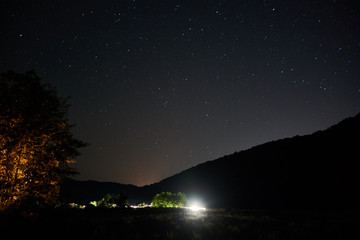 Night starry sky over the mountains. Stars and constellations are visible through light clouds. The bright light of the headlights of a car in the distance.
