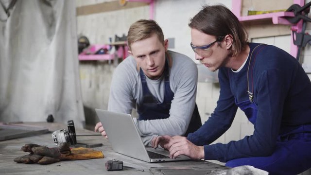 Side view tracking shot of two young metalworkers discussing project on laptop computer screen standing at workbench surrounded by tools