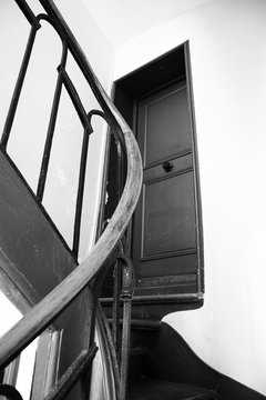 Typical narrow spiral staircase with railing in old Parisian house and flat door. Selective focus. Black and white photo,
