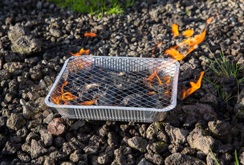 Small disposable barbecue stands on the stones. Flames come from charcoals of grill. Preparing BBQ,...