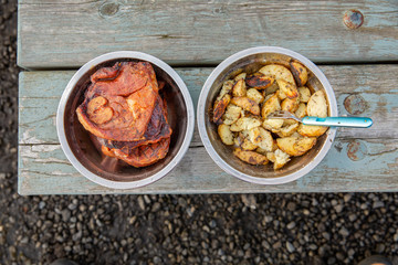 Metal bowls with lamb steaks and potatoes on wooden picnic table. Top view.