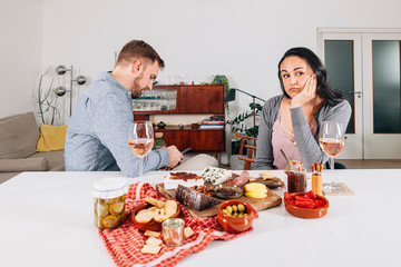 a clearly bored woman sits in front of a table laden with food while her partner does not pay attention to her because he is busy on his mobile phone
